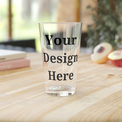 Single clear pint glass featuring “your design here” sitting on wooden counter.