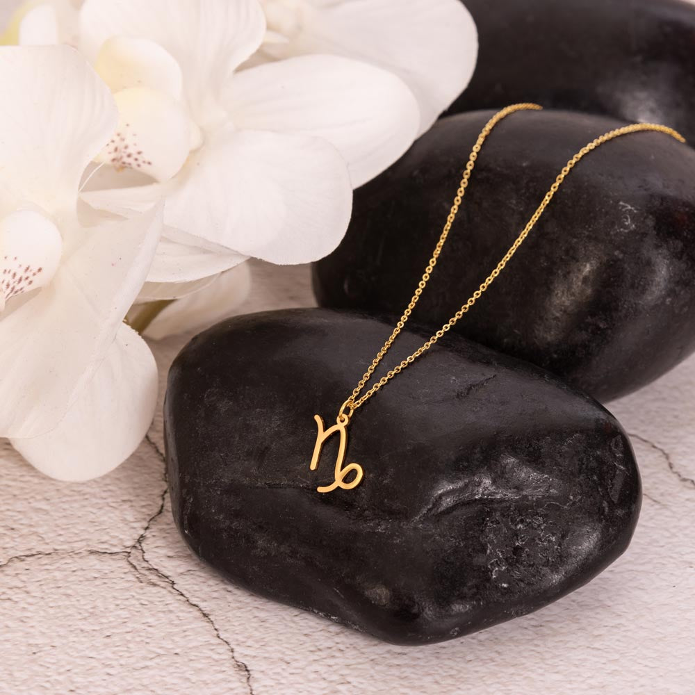 Gold zodiac charm necklace displayed on polished black rock.