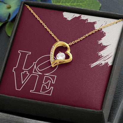 Polished gold heart necklace hanging in front of LOVE message card in standard black jewelry box.